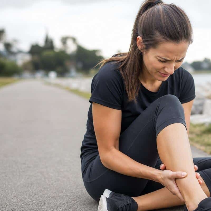 Woman in athletic wear sitting on a lakeside path, grimacing in pain while clutching her lower leg and Achilles tendon area.